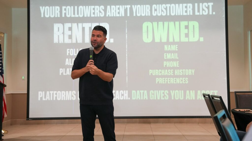 A man in a black polo shirt speaking into a microphone before a presentation