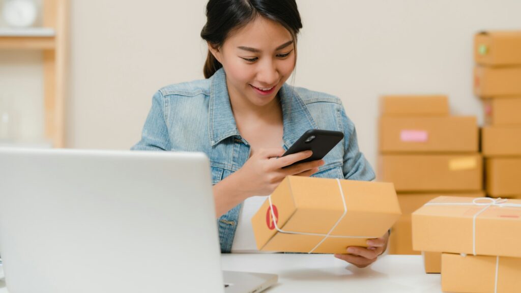 Woman checking package with phone near laptop and boxes