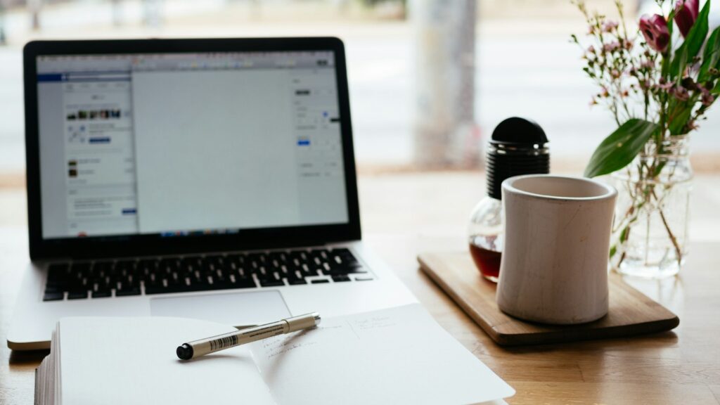 Laptop, open notebook and pen on a wooden desk beside a mug