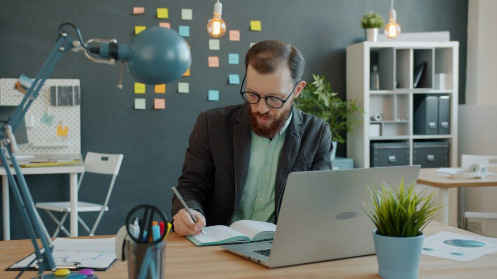 Man writing in a notebook at a desk with a laptop