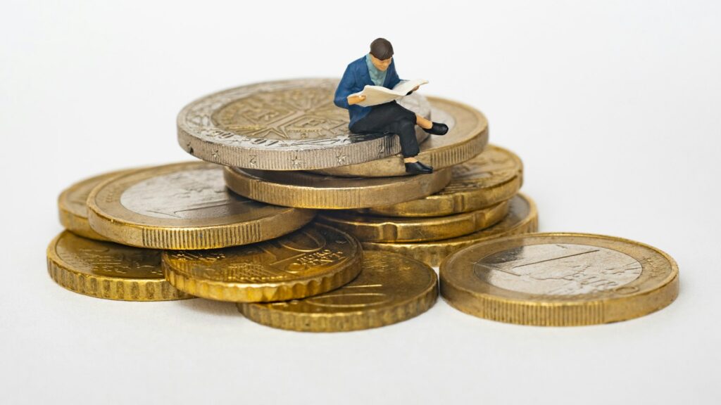 Miniature person sitting on stack of coins reading newspaper
