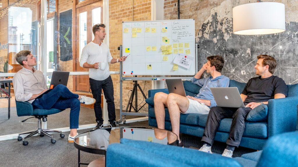 Three men sitting while using laptops and watching man beside whiteboard